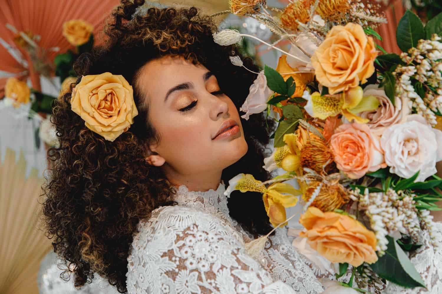 A bride in a high neck lace gown wears a yellow rose in her hair and holds a bouquet with various orange and coral colored roses.