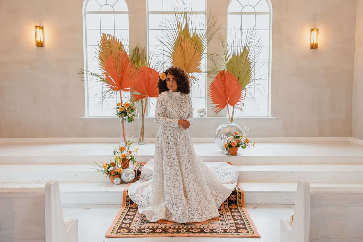 A bride in a high neck lace gown poses in front of large windows decorated with yellow and orange flowers.