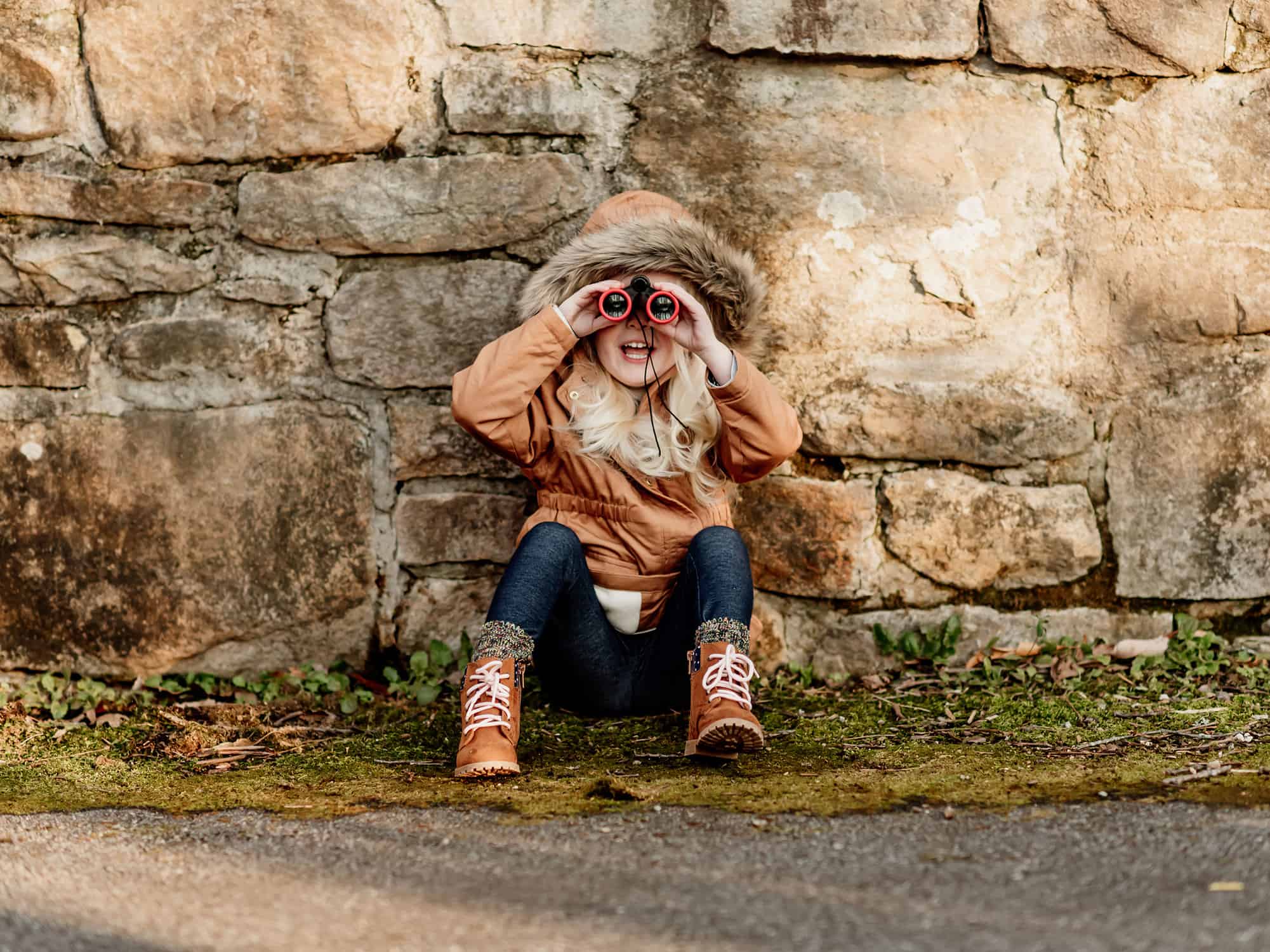 Young child sitting against a wall outside looking through binoculars